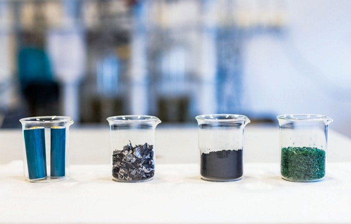 A set of four beakers: the leftmost one shows two blue batteries, the left center shows black chunks of batteries, the right center shows a black powder, and the rightmost one shows shredded green material.