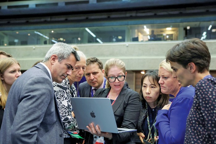 During the third UN meeting on plastic pollution, a group of men and women crowd around a woman with blond hair and glasses holding an open Apple laptop.