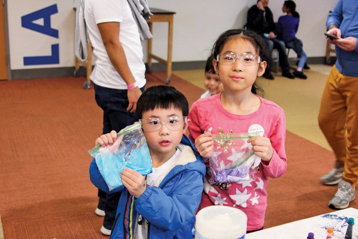 Two children wearing safety glasses display plastic bags filled with colored slime.