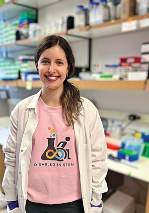 A scientist standing in a biology lab while smiling at the camera. She’s wearing a lab coat over a T-shirt with the words “Disabled In STEM” written on it.