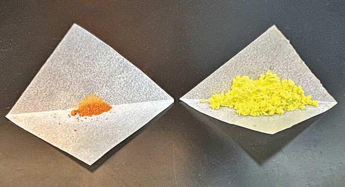Two piles of powder on pieces of white weighting paper on a dark-colored benchtop. The pile on the left is orange and much smaller than the pile on the right, which is yellow.