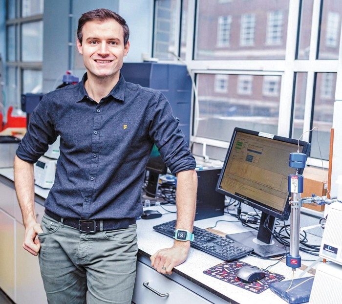 A photo of Stephen Cochrane standing and smiling. He is wearing a dark blue button-up shirt and gray pants.