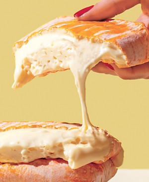 Photo of Minger cheese, a melty white cheese with a thick rind shown cut in half and held above a plate in a person's hand.