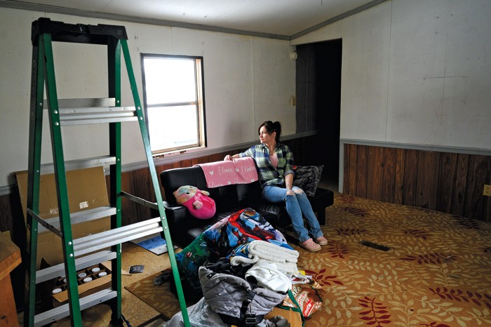 A woman sits on a couch in a trailer and looks out a sun-filled window. A ladder and pile of clothes are in the foreground.