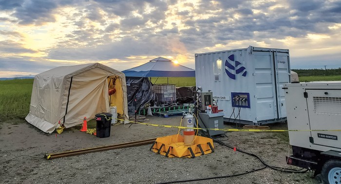 A generator is hooked up to a small shipping container that is receiving water from a tote under a pop-up canopy next to a tent.