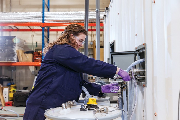 A scientist collects clean water in a beaker from a faucet on the side of a shipping container that houses a supercritical water oxidation unit for destroying per- and polyfluoroalkyl substances.