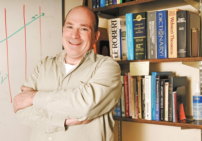 A photo of David Saltzberg standing in front of a bookshelf and whiteboard.