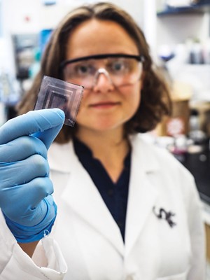A researcher wearing safety goggles holds a square piece of glass patterned with a semitransparent material in front of her with gloved hands.