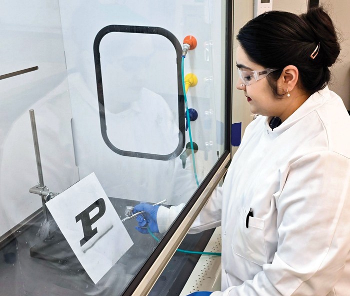 A researcher uses a spray gun to create an underlined letter P on a piece of paper placed behind a fume hood door.