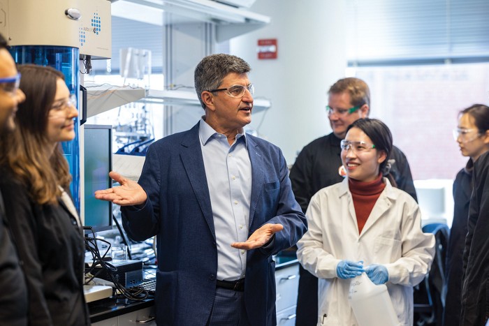 A group of people standing and talking in a laboratory. The person in the center gestures to his left.