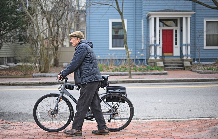 Dan Hogan walks a bicycle along a brick sidewalk in Harvard Square. He wears a dark winter coat and a flat cap.