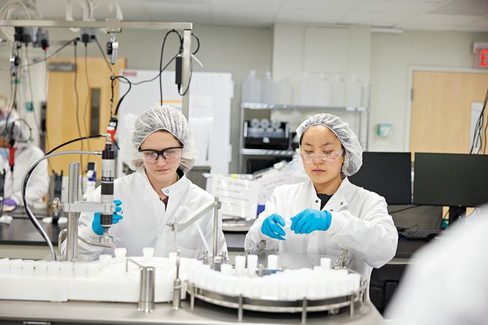 Two technicians, each wearing blue nitrile gloves, safety glasses, a lab coat, and a hair net, process samples in a laboratory.