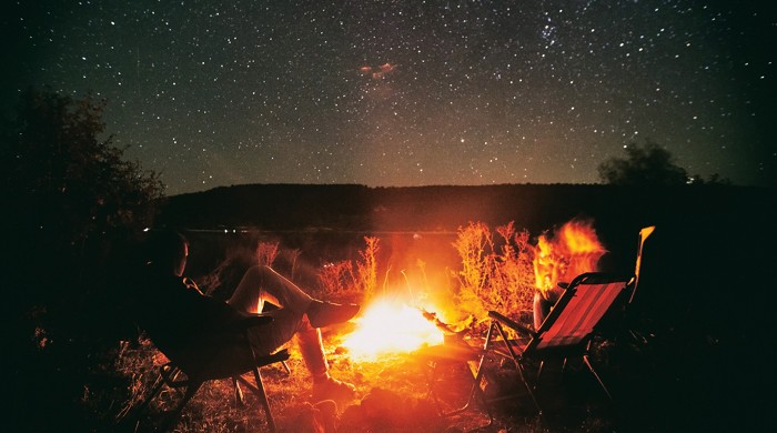 People are sitting on chairs in front of the fireplace. Above the orange glow of the fire is a distant ridge, above which many stars dot the sky.