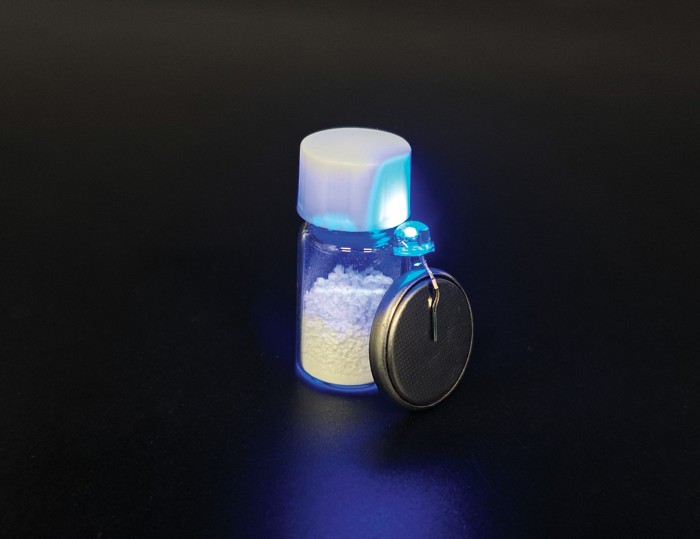 A glowing blue light-emitting diode attached to a coin-cell battery resting on a small glass bottle filled with white powder.