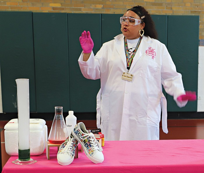 Woman in laboratory protective clothing standing and talking behind a table that holds lab equipment and sneakers.