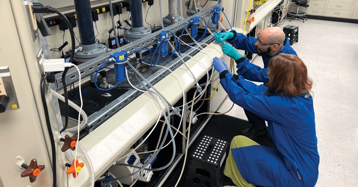 Two scientists in blue coats manipulate tubing inside a chemistry lab.