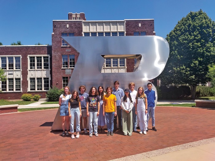 A group of eleven smiling students stand in front of a large silver statue in the shape of a capital letter B.