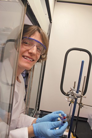 A smiling student wearing safety glasses, a lab coat, and gloves leans into a fume cupboard.