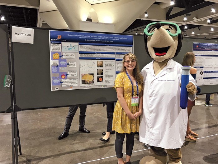 A smiling student stands in front of a poster alongside a large mascot mole wearing a lab coat.