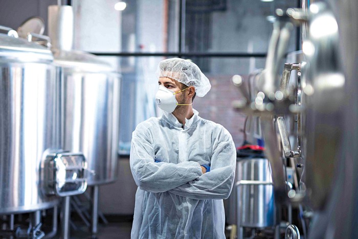 A worker at XChem, who is standing next to two small reactors and wearing a face mask and head covering.