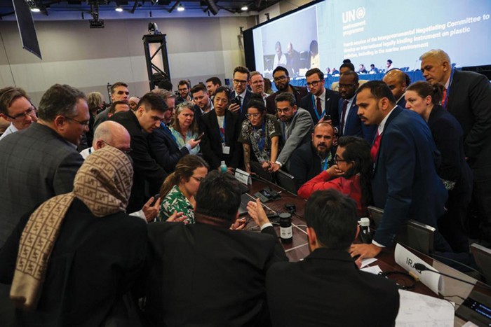 A group of people huddled around a woman seated at a long table, trying to reach a compromise on a UN treaty document.