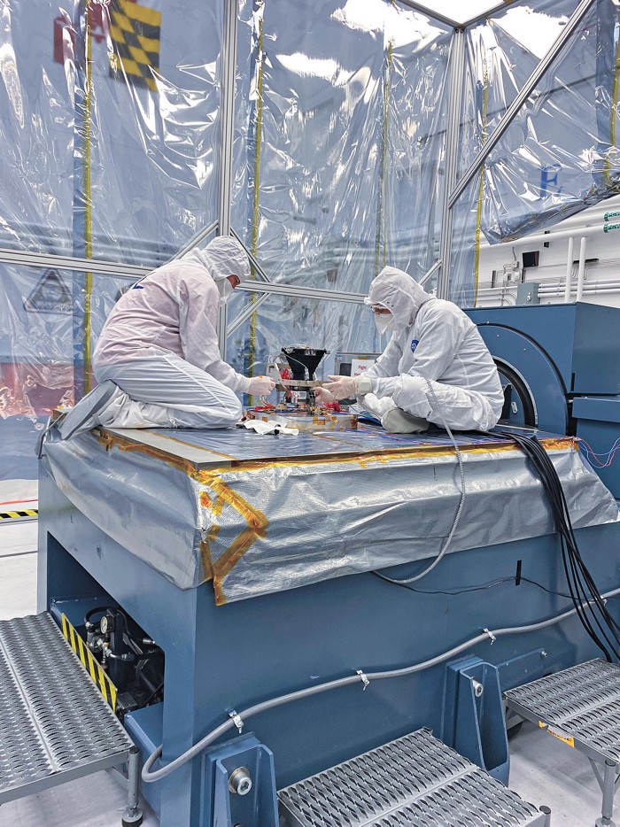 Two scientists garbed in full-body cloth coveralls, gloves, and masks sit on a large table adjusting a small instrument between them.