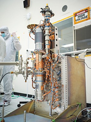 A silver tube with copper wires is bolted vertically to a metal box. Researchers in full cloth coveralls, gloves, and face masks stand talking in the background.