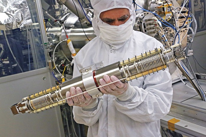 A man in full-body cloth coveralls, a face mask, and gloves holds a long, silver tube.