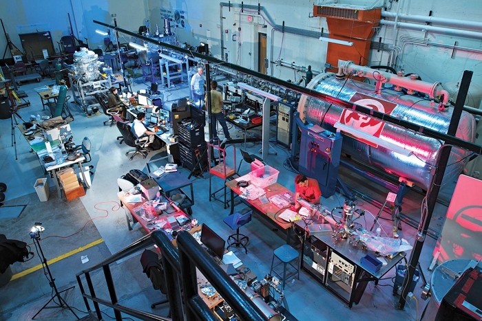 A warehouse-like lab space holds work benches and a large, cylindrical dust accelerator.