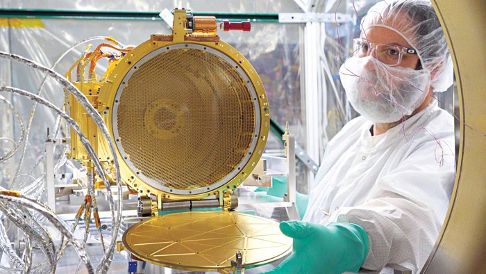 A researcher garbed in cloth coveralls, gloves, and hair and beard nets opens a wide, golden cylinder toward the camera.
