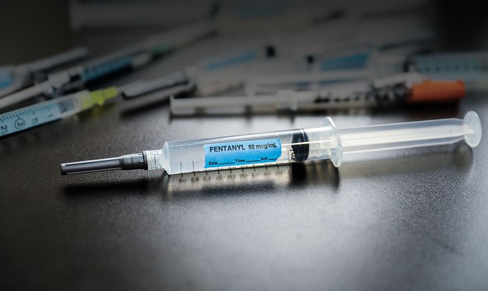 A syringe sits on top of a table. It has a blue label marked with black ink spelling out "fentanyl." Several additional syringes are in the background.