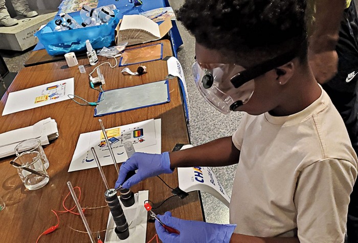 A child in goggles and gloves stands in front of a table with a voltaic pile on it.