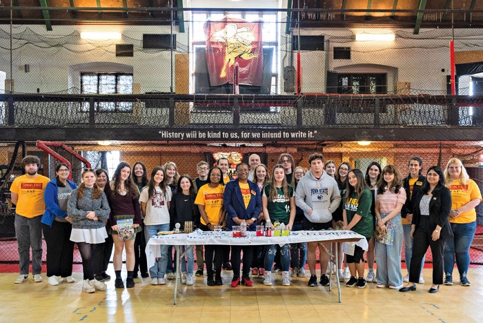 Volunteers stand in a large group behind a table that holds equipment for a battery demonstration.