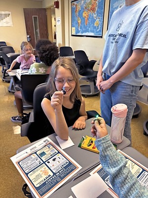 A smiling child sits at a desk and holds a button battery and a light-emitting diode bulb.