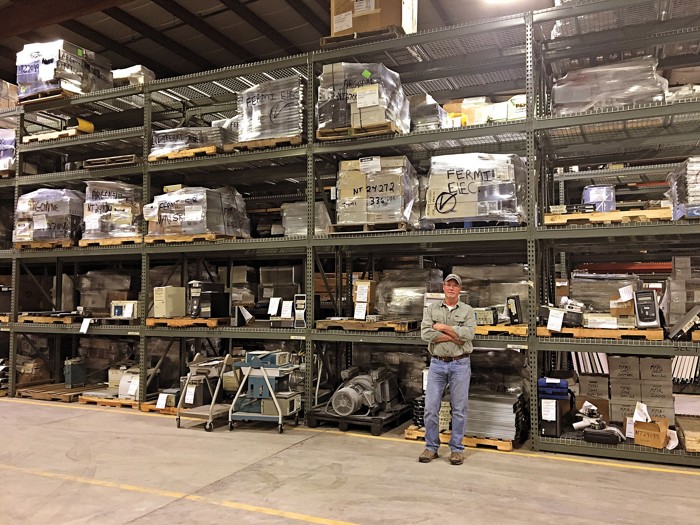 A man in blue jeans and work boots stands in front of a shelving unit in a warehouse. It makes him look small. Some nearby pallets hold recognizable scientific equipment, others are wrapped in plastic and labeled "Fermi."