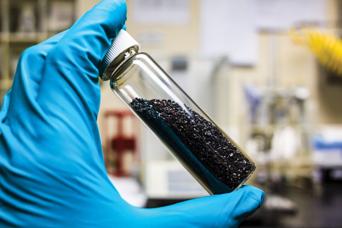 A gloved hand holds a small glass vial containing granular activated carbon.
