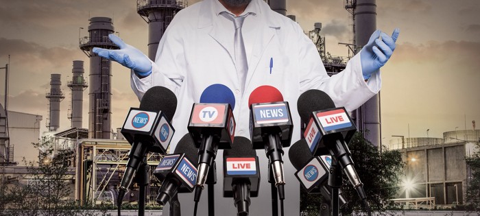 A person in a lab coat stands in front of several microphones.  They are wearing blue gloves and a silver tie.  Behind them is a chemical factory.