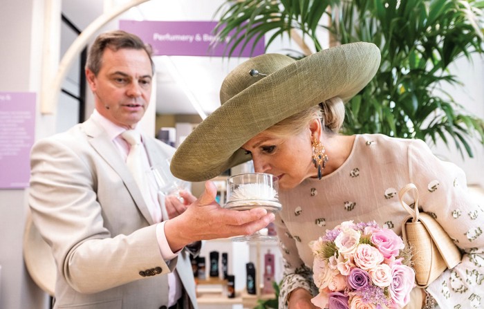 Queen Máxima of the Netherlands, wearing an olive-colored straw hat, smells a flower held out for her in a glass container.