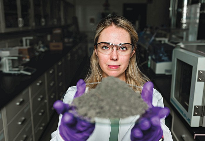A scientist looks at the camera while holding a plate with a pile of gray ash.