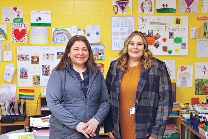 Two women stand side by side in front of a wall covered in children's drawings