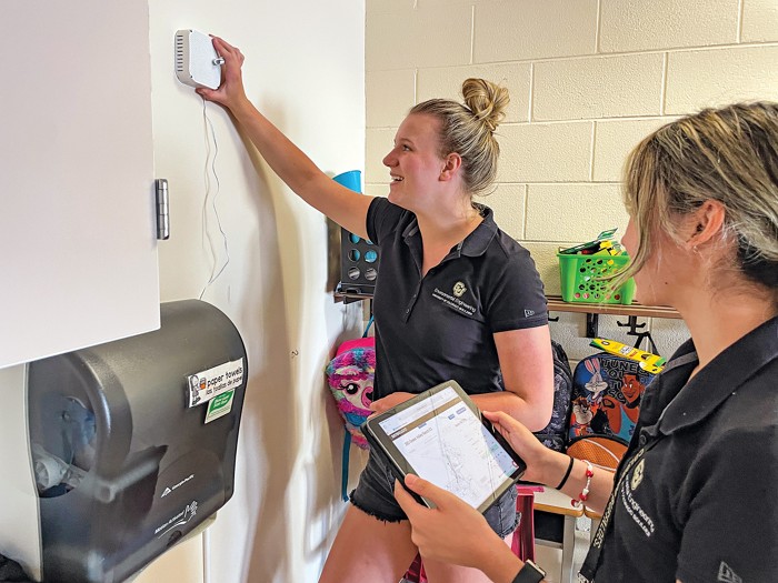 Two people stand in front of a wall. One is reaching up and holding a small white box while the other looks at a hand-held tablet.