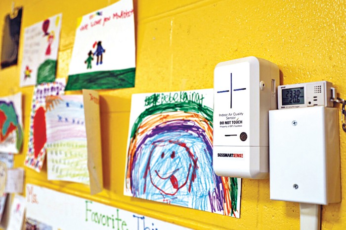 A white box with "Indoor Air Quality Sensor DO NOT TOUCH" printed on it is attached to a yellow wall. To the right are many children's drawings