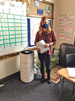 A teacher in a mask stands in her classroom next to a waist-high white tower.