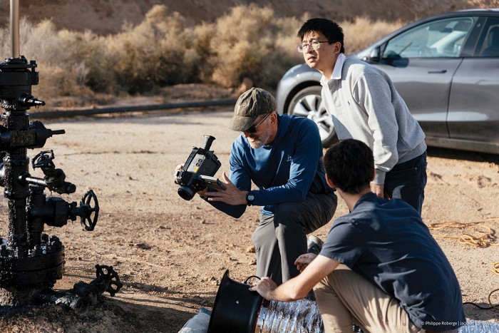 Rob Jackson and two colleagues crouching on a dusty ground in scrubby terrain. They’re next to a pipe sticking out of the ground with several valves sticking out of it at various angles. Jackson is holding a methane sensing device that looks like a camcorder.