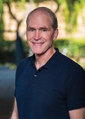 Rob Jackson, smiling, wearing a dark blue short sleeve polo shirt. In the background, trees and greenery can be seen out of focus.