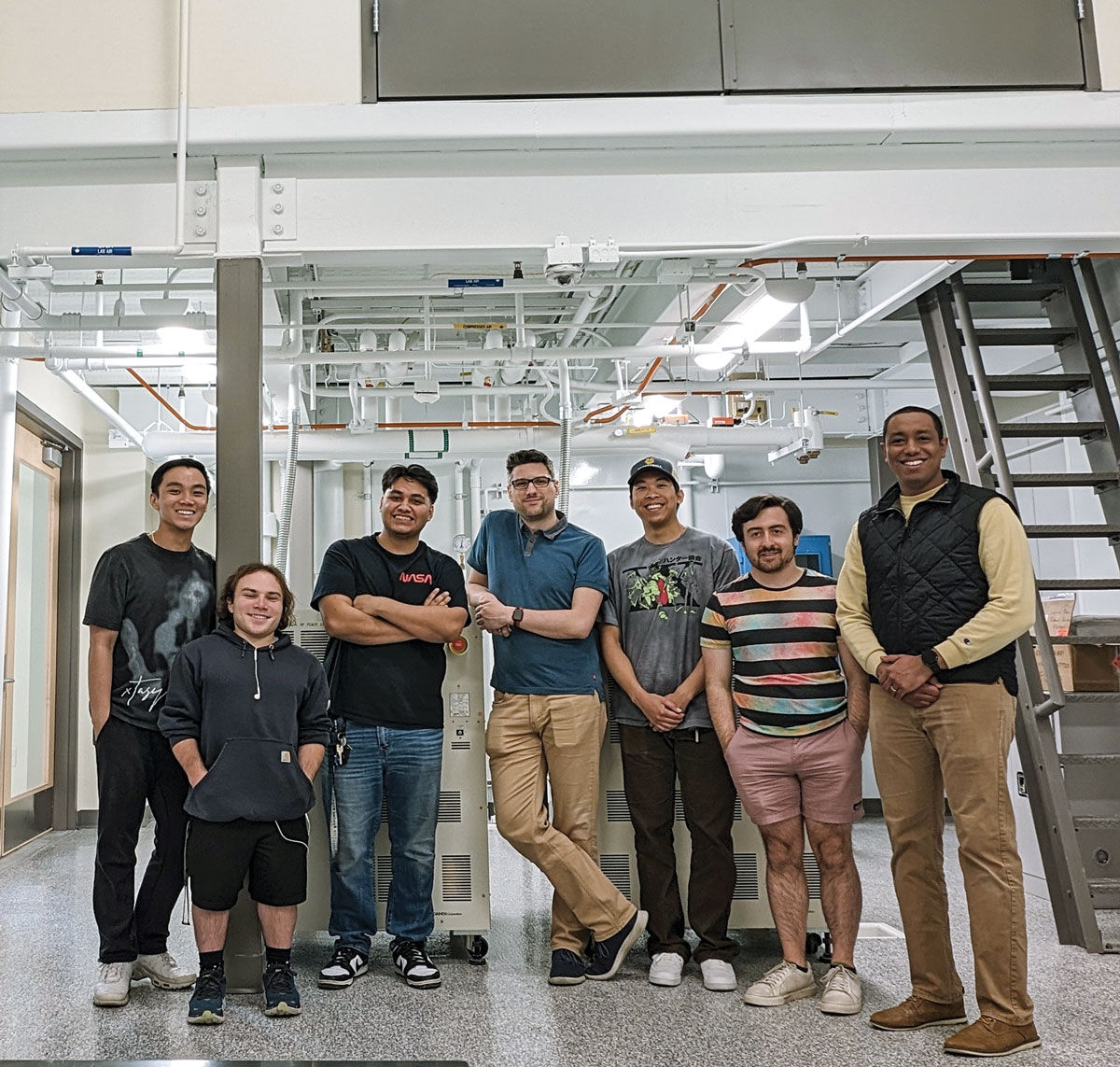 Seven people stand in front of a brightly lit space with lots of white pipes running along the ceiling and a ladder to the far right.