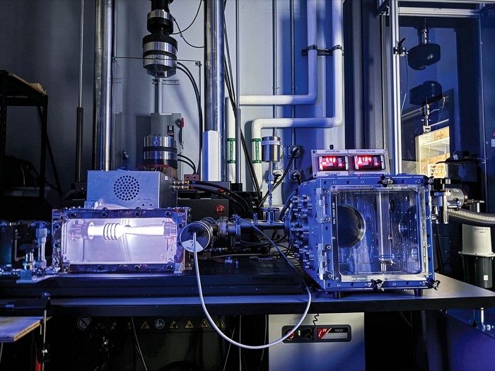 A lab bench holds two boxes that sit side by side and are connected by wiring, The one on the left glows white, and the box on the right has red dots of light on top. Pipes run against a wall in the background.