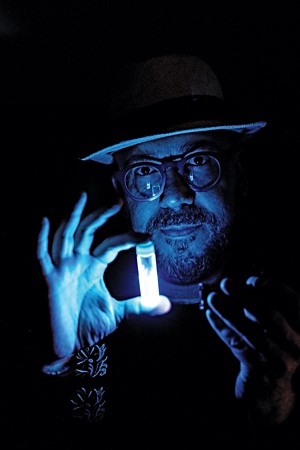 Alán Aspuru-Guzik is in a dark room holding a vial that contains a substance that is glowing bright blue.