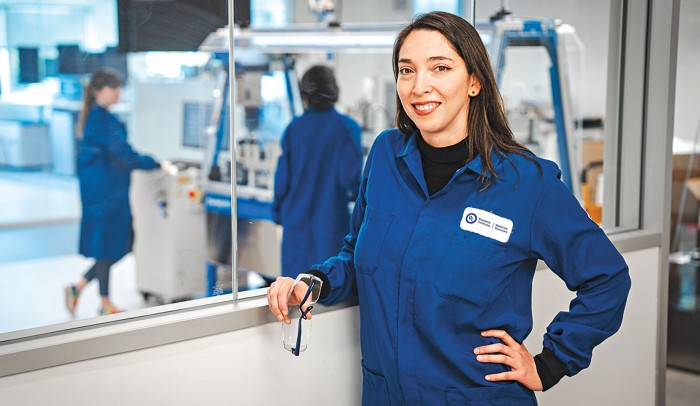 Varinia Bernales wears blue lab coat and leans against a wall with a window. Two researchers are reflected in the window behind Bernales.