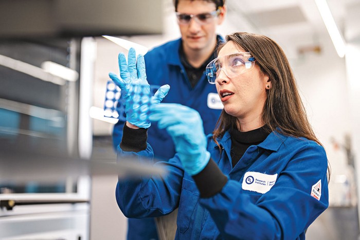 Varinia Bernales holds a 3D-printed vial holder and is wearing a blue lab coat and blue gloves. Conor Brew stands slightly behind, looking at the vial holder. 